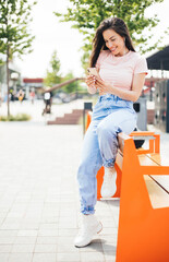 Beautiful cheerful smiling woman with earphones listening music on her mobile phone sitting outdoors at the street while enjoying.