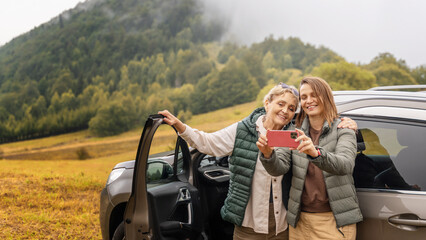Two happy adult women senior mother and daughter traveling together by car taking selfie standing...