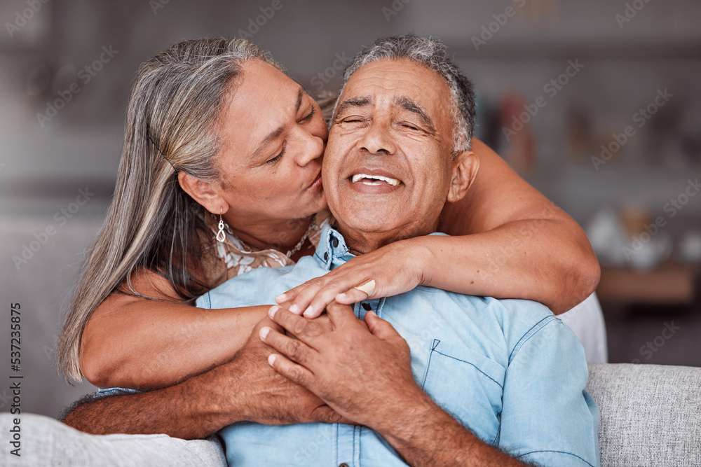 Poster Happy senior couple hug in home, retirement lifestyle together in Mexico and relax on sofa. Romantic marriage life, love bonding time and elderly woman kissing latino husband on cheek in living room