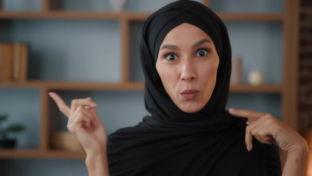 Happy ecstatic exited active Arabian Muslim Islamic 30s woman in black hijab celebrate win victory at home having fun indoors with funny dace to music dancing with hands smiling looking at camera