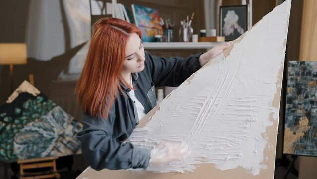 Red-haired Caucasian woman girl lady artist painter decorator at home in art studio creates picture on canvas spreads white thick paint with hand uses putty to create texture hobby painting with hands