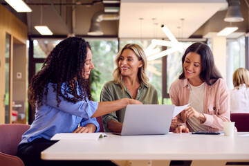 Multi-Cultural Female Business Team Sitting At Desk In Open Plan Office Collaborating On Project