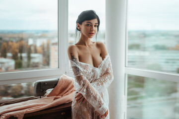 Sexy girl in a white translucent tunic sits sewing near the window.Beautiful female body shines...