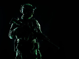 Fototapeta na wymiar Army special forces elite soldier with hidden behind mask and glasses face, battle helmet, tactical radio headset, standing with assault rifle equipped silencer in darkness, contour shot