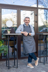 Fototapeta na wymiar Mature man in plaid shirt and apron standing at the cafe entrance