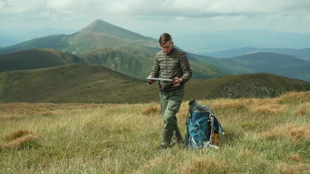 A man takes out a laptop from a backpack and sits down to work, outdoors in the mountains. Remote work with a view of the mountains. Freelance in nature on a sunny day. High quality 4k footage