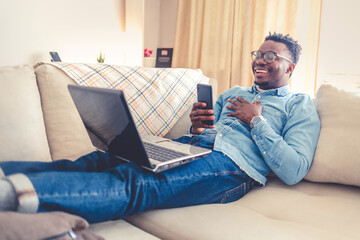 Smiling young 30s man sitting relaxed on sofa in living room browsing internet on modern computer...