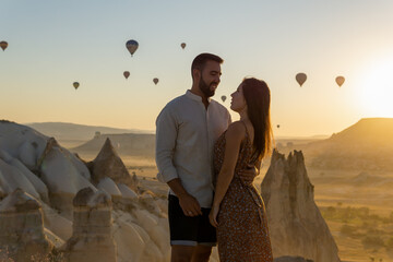 Young couple looking into each other's eyes in the foreground watching the typical hot air balloons of the cappadocia area flying at sunrise.