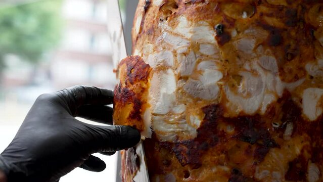 A cook is cutting delicious meat with a big sharp knife to make shawarma d&ouml;ner kebab in black gloves. Cooking chicken meat on a vertical skewer. Bright and slow motion 4k video in grill restaurant.