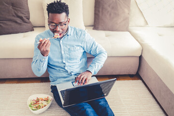 Young man relaxing at home, sitting on the floor eating a takeaway meal while working on the laptop at home during the day.