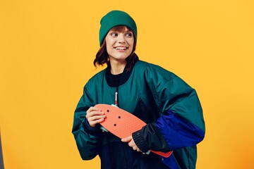a beautiful, stylish woman stands in a jacket and hat on a yellow background holding a small skateboard in her hand and smiling happily