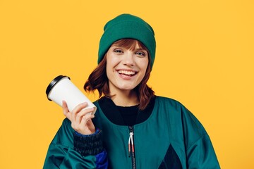 a happy, joyful woman stands on a yellow background in a stylish jacket and holding a coffee glass in her hands, smiling broadly and leaning the glass against her face. Horizontal Studio Photography