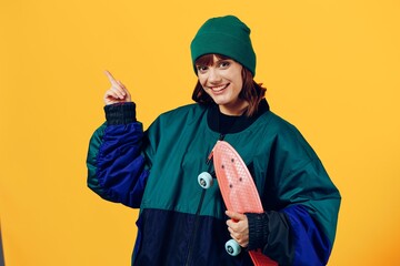 a beautiful, stylish woman stands in a jacket and hat on a yellow background holding a small skateboard in her hand and with her free hand points to the empty space of the background