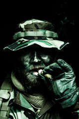 Brutal and serious commando soldier, army special forces veteran, in camouflage battle uniform, boonie hat, black paint on bearded face, combat knife in shoulder holder, smoking cigar, studio portrait