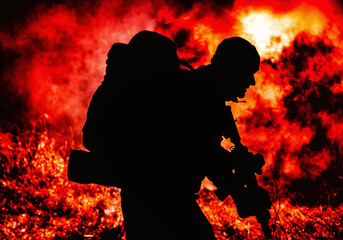 Silhouette of army soldier, elite troops fighter armed service rifle, carrying tactical backpack on back, standing on background of fiery exposure. Combatant moving in darkness on burning battlefield