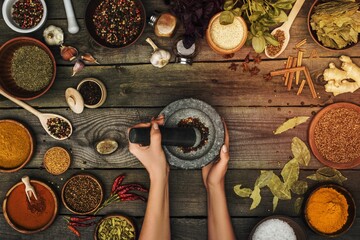 Top view of a woman grinding pepper with pestle in a mortar and various spices on a wooden table - obrazy, fototapety, plakaty