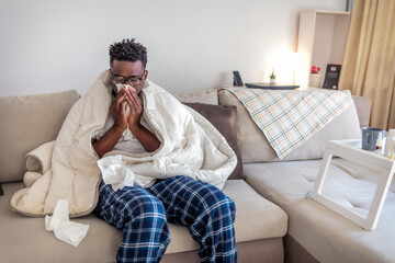 Cropped shot of a young man suffering with flu while sitting wrapped in a blanket on the sofa at home.