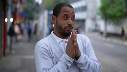 One anxious black man standing in urban street feeling nervous and stressed. Desperate African American rubbing hands in anxiety in modern sidewalk