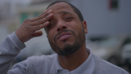 Obraz premium Frustrated young man feeling angst rubbing forehead with hand standing outdoors in street. One anxious black African American suffering