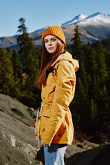 Woman in yellow raincoat with red hair on a hike stands in front of mountains in yellow cap autumn sunset light 