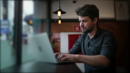 Concentrated young entrepreneur working in front of laptop computer by window in urban city cafe