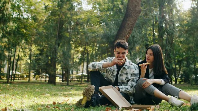 Happy Couple Eating Pizza In The Park And Talking. Attractive Young Man And Woman Sitting On The Grass On A Bright Sunny Day, Having A Picnic, Dating, Enjoying Fast Food. Takeout, Delivery Concept