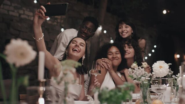 Ethnically diverse group of friends taking selfie with young lesbian newlyweds during wedding party in modern restaurant
