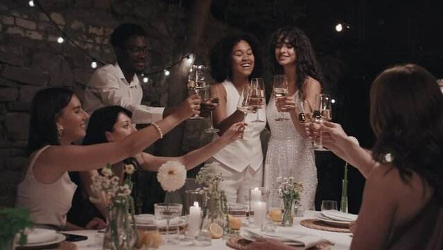 Modern lesbian newlyweds, their relatives and friends clinking glasses with champagne or sparkling wine at wedding party in evening