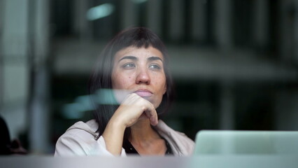 Obraz premium Pensive young woman thinking while working in front of laptop seen through window reflecting city sidewalk street. One thoughtful hispanic female businesswoman