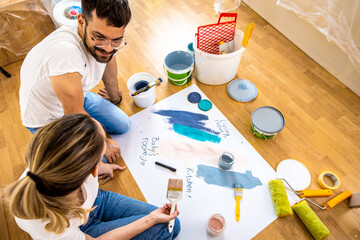 Young couple sitting on the floor choosing color for painting the wall in their home.	