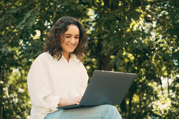Photo of smiling young woman with curly hair sitting on bench and working on laptop outdoor.