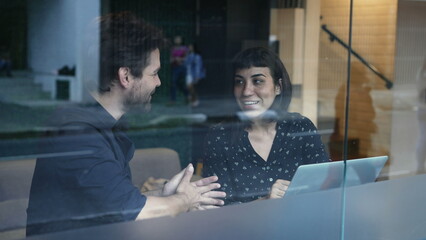 Fototapeta na wymiar Two young people talking about work in front of laptop and workplace seen through window reflection. Business woman and man discussing in conversation