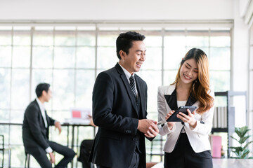 Senior Asian businessman and young female businesswoman discuss in the office, working on social media strategy, looking at a tablet with blur colleagues in the background. Image with copy space.