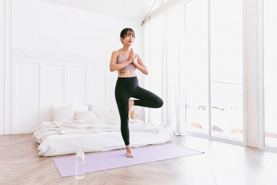 Beautiful happy young Asian woman standing in Vrksasana Posture, doing yoga exercise on purple mat with water bottle, working out in bedroom, indoor workout, Tree Pose, full length image.
