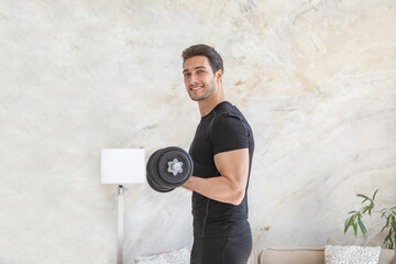Side view of young handsome man in black sportswear doing biceps curl with dumbbells in living room...