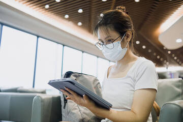 A white Asian woman wearing a mask in public, using a digital tablet while waiting for a flight at the airport. Business travel concept.