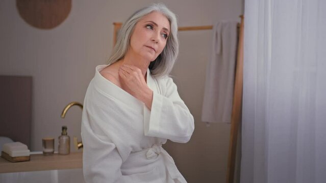Old elderly 60s Caucasian woman in bathrobe in bathroom smears shoulders back and neck with lotion body cream vitamins collagen uses oil for soft skin rejuvenation anti-wrinkle procedure moisturizing