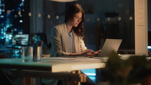 Office at Night: Focused Female Manager Working Long Hours Alone in Creative Agency, Implementing Business Strategy for Client. Creative Specialist Writing Corporate Project Plans on Laptop Computer.