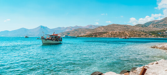 Gulf of Elounda. Pleasure boat in the bay against a mountains ridge and blue cloudless sky. The view of the pleasure boat in Elounda bay from the Spinalonga island - obrazy, fototapety, plakaty