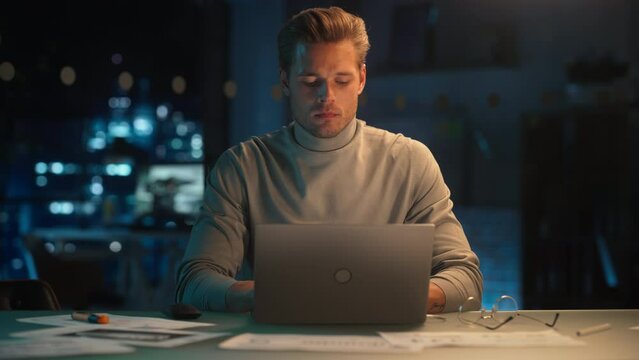 Handsome Man Working on Laptop Computer in a Company Office at Night. Young Specialist Analyzing Reports. Marketing Assistant Developing a Consumer Sales Strategy for a Client.