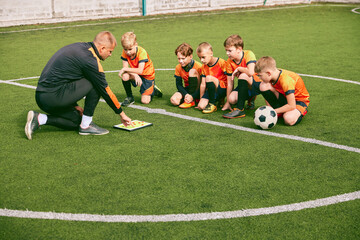 Football training. Soccer coach explaining game rules and strategy using tablet, map. Sports junior team sitting on grass pitch with trainer