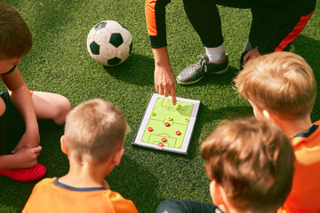 Football training. Soccer coach explaining game rules and strategy using tablet, map. Sports junior team sitting on grass pitch with trainer