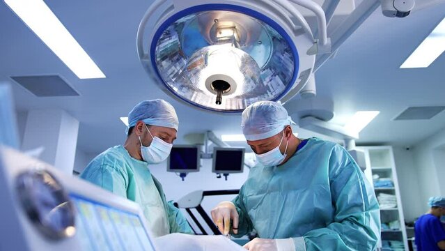 Male doctors performing operation under a big lamp in surgical room. Surgeons uses medical instrument device producing little smoke.