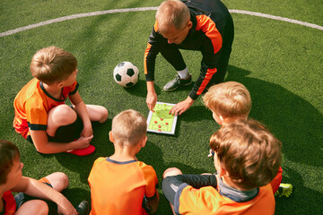 Football training. Soccer coach explaining game rules and strategy using tablet, map. Sports junior team sitting on grass pitch with trainer