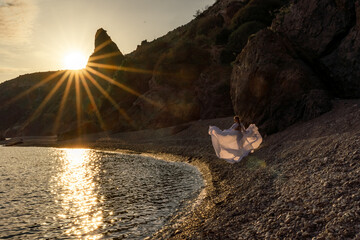 A mysterious female silhouette with long braids stands on the sea beach with mountain views, Sunset rays shine on a woman. Throws up a long white dress, a divine sunset.