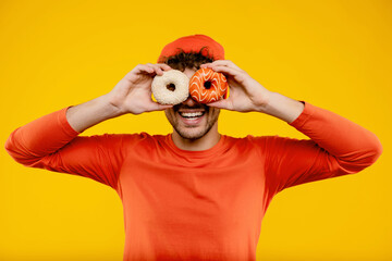 Young handsome smiling man holds donuts in both hands like glasses on isolated yellow background