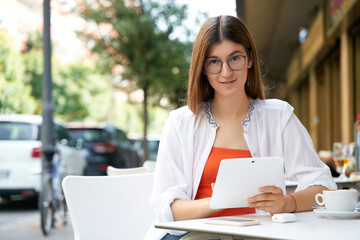 Smart student girl in eyeglasses sitting with digital tablet and cup of coffee during language class in cafe terrace. Remote job concept 