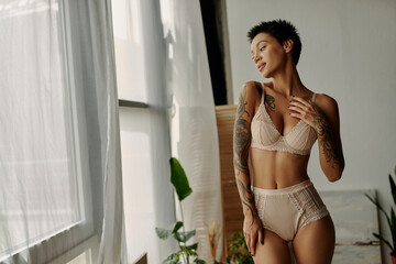 Young tattooed woman in lace lingerie looking at window at home.