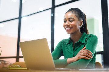Smiling young african-american business woman wearing casual shirt using laptop for office work, female employee looks at the screen, reading news, checks mail