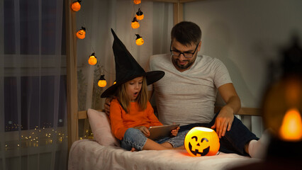 Father and little child in witch black hat watch horror film on tablet in dark with orange pumpkin lantern. Halloween celebration